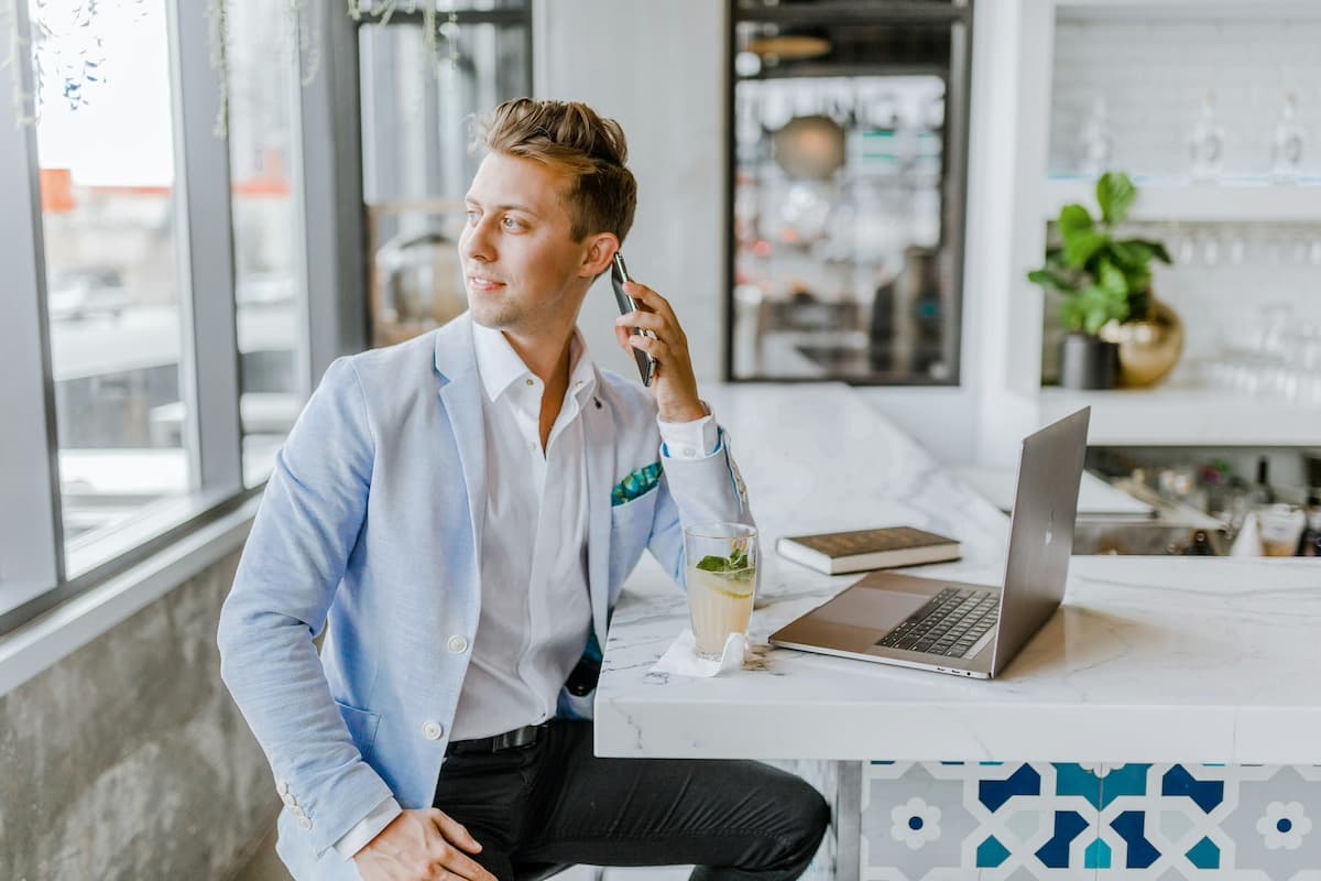 Business man in a bleisure travel sitting at a bar with his phone to his ear and a laptop and cocktail on the counter