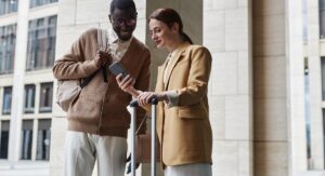 Young businesswoman with suitcase consulting with black man about way to airport while showing him a travel management software