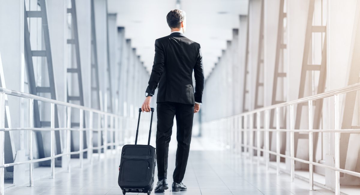 Rear view of tall businessman in stylish suit walking by airport terminal, carrying luggage