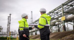 Two oil and gas workers in high-visibility jackets and hard hats reviewing plans at a refinery site