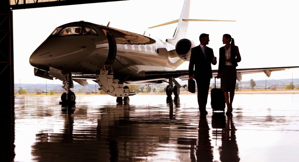 A man and a woman, both adults, stroll near a private jet charter inside an airplane hangar, the man holding a briefcase and the woman rolling a suitcase.