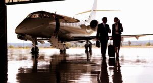 A man and a woman, both adults, stroll near a private jet charter inside an airplane hangar, the man holding a briefcase and the woman rolling a suitcase.