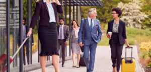 everal business professionals commute to work on a street, showcasing holiday executive travel. A woman in a dark blazer and skirt is visible on the left, pulling a suitcase. Two men and two women are behind her; one woman is talking on her phone, while a man with grey hair is carrying a briefcase.