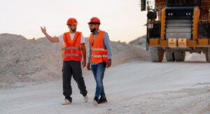 Two adults wearing orange vests and hard hats are walking along a dirt road in front of a large yellow truck, with one pointing. They are wearing safety vests and work attire. The setting appears to be a construction site